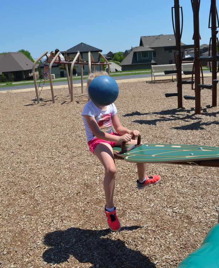 Child on seesaw at playground with ball perfectly covering face, a funny and interesting picture taken at the perfect time.