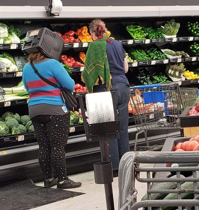 Shoppers in Walmart produce aisle, one wearing a plastic bin on their head, highlighting unusual store moments.