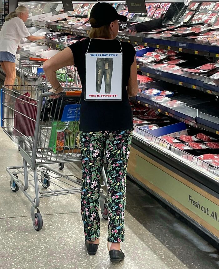 Woman shopping at Walmart meat aisle wearing floral pants and shirt with anti-ripped jeans message, unusual store scene.