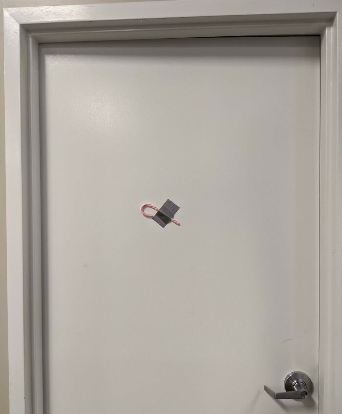 Minimalist workplace Christmas decoration showing a single candy cane taped to a plain white door with duct tape.