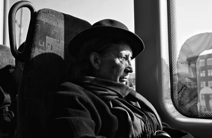 Elderly woman wearing a hat and scarf, sitting by a window capturing street moments that feel like magic in daily life.