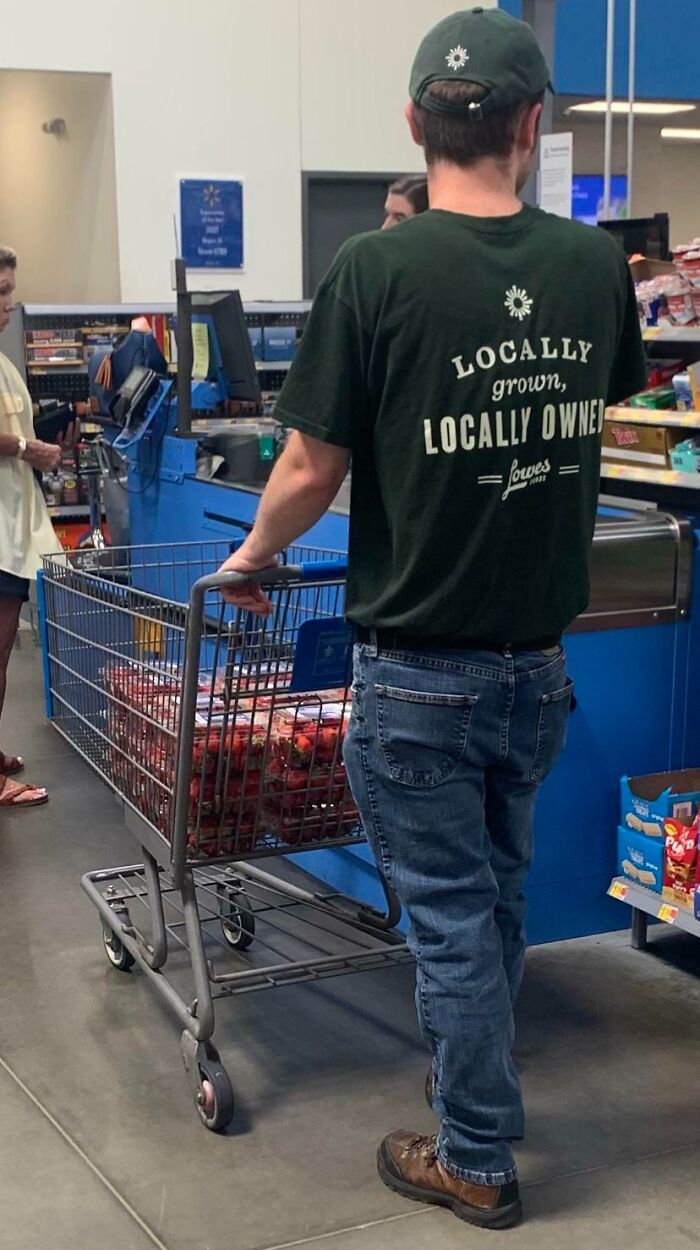 Man in Walmart wearing green shirt with locally grown logo, pushing cart filled with produce at checkout lane.