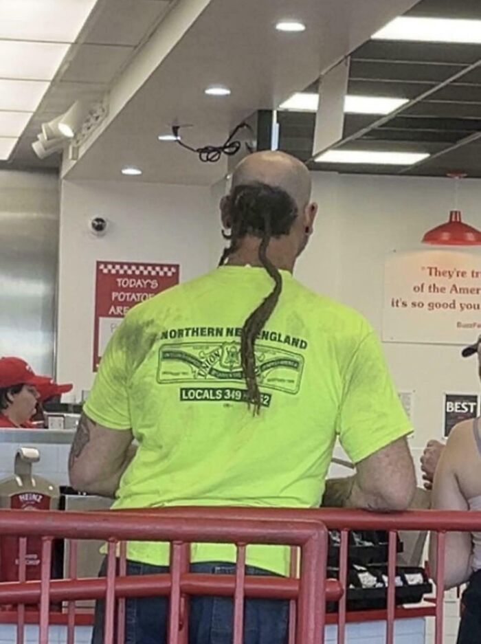 Man with unusual braided hairstyle and bright yellow shirt standing inside a Walmart store with other customers nearby.
