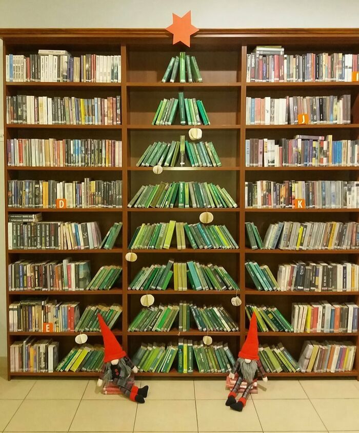 Books arranged as a Christmas tree in a workplace bookshelf, featuring creative Christmas decorations and two elf dolls.