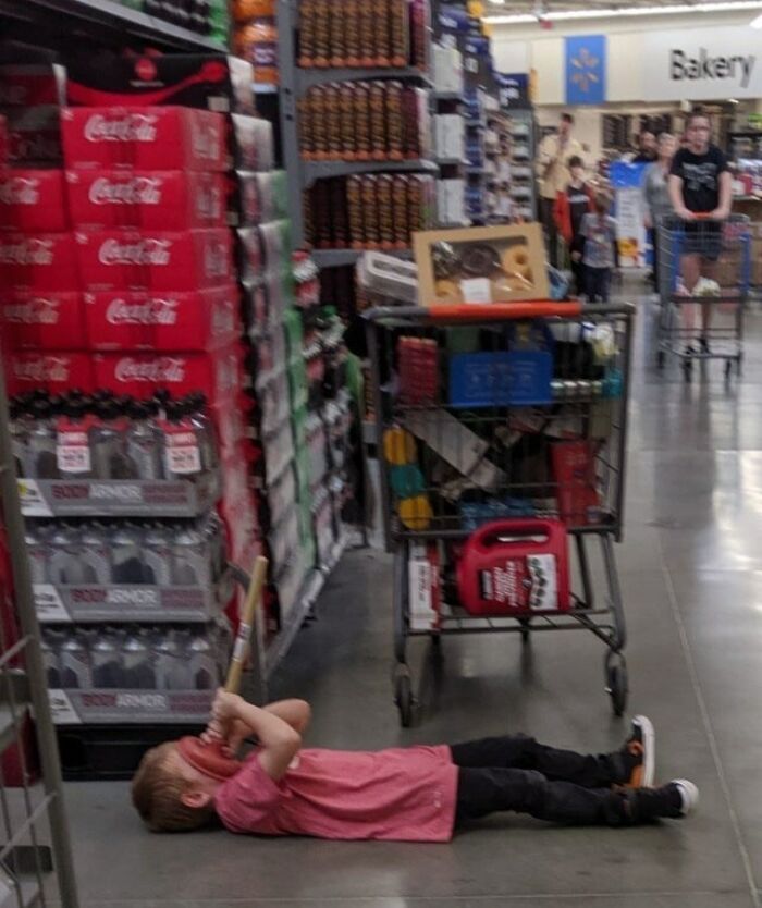 Child lying on the floor in a Walmart aisle near stocked shelves, with shopping carts and customers in the background.