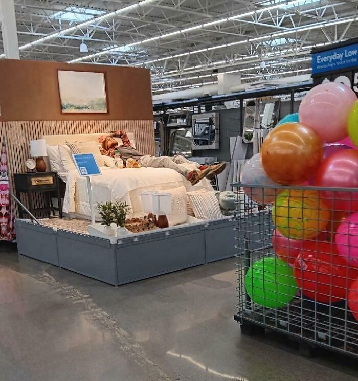 Person lying on a bed display inside a Walmart store with colorful balls in a metal bin nearby, unusual Walmart scene.