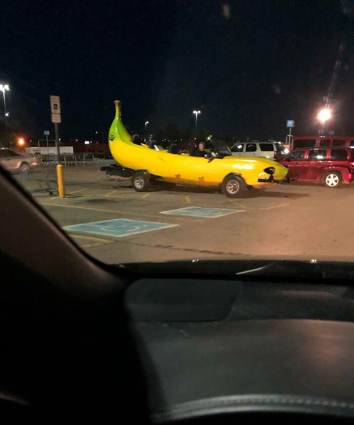 Unusual banana-shaped car parked at night in a Walmart parking lot, drawing attention from shoppers nearby.