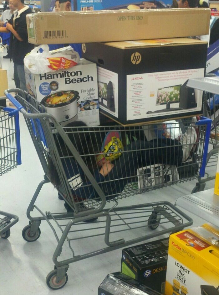 Shopping cart at Walmart filled with electronics, kitchen appliances, and a person lying inside the cart amidst unusual store items.
