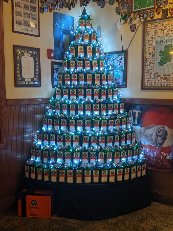 Creative workplace Christmas decoration using stacked Jägermeister bottles arranged as a tree with blue lights in a pub setting.