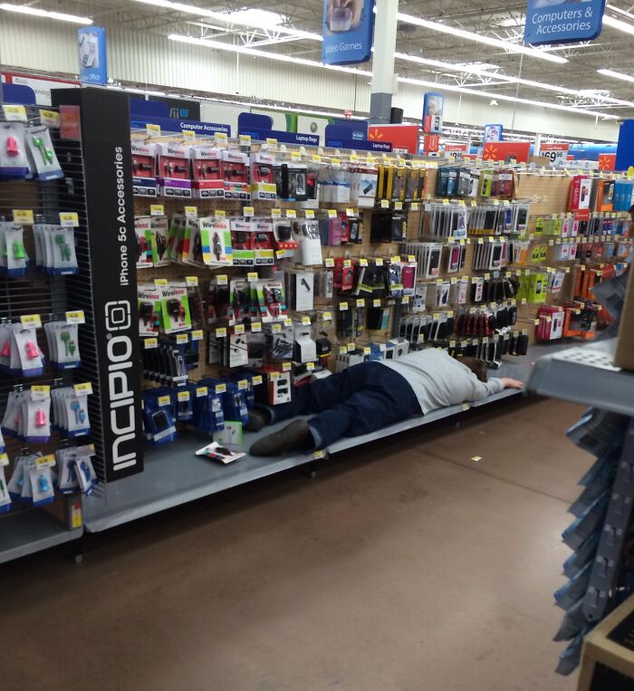 Person lying on display shelf in Walmart electronics aisle surrounded by phone accessories in a large store.
