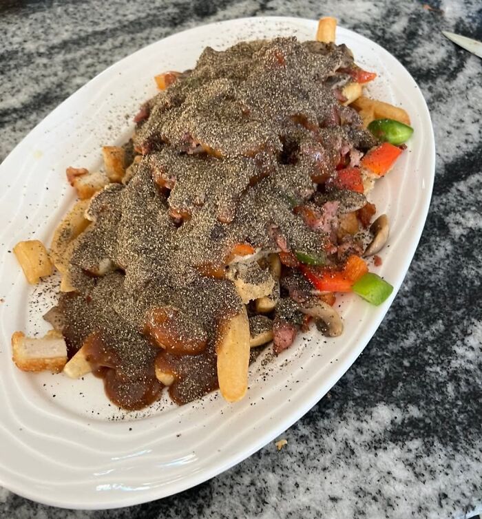 Plate of fries covered in brown gravy, vegetables, and an excessive amount of black pepper, a food pic to be shamed online