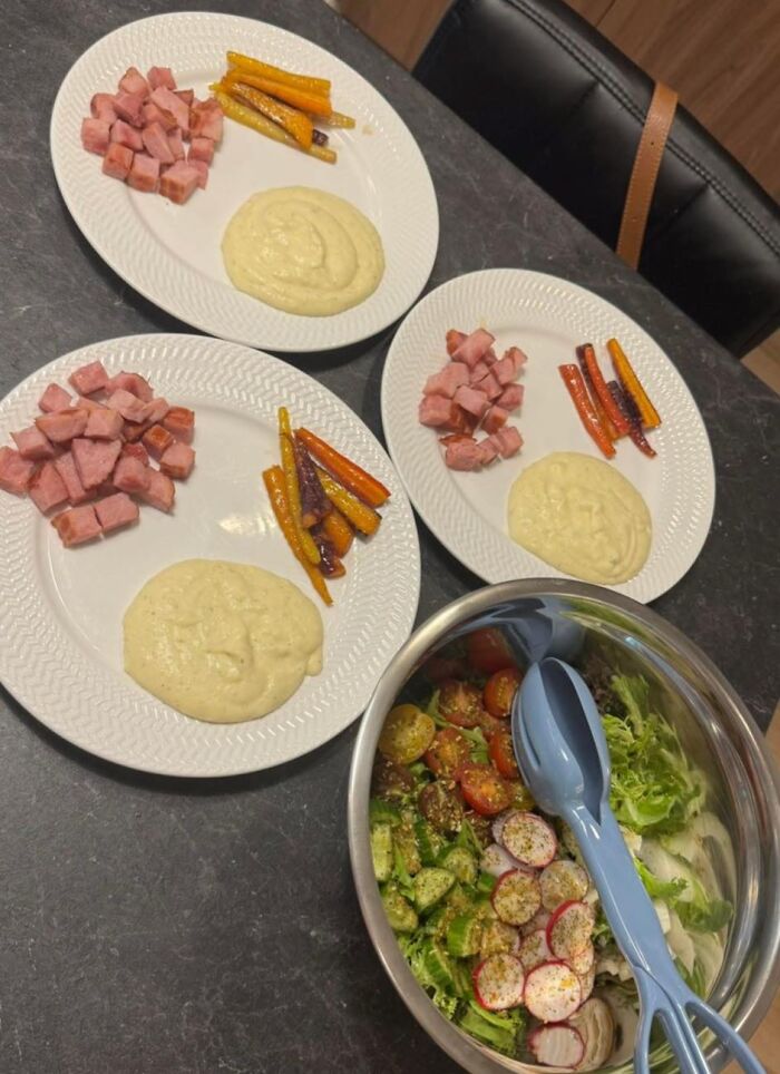 Three plates with cubed ham, roasted carrots, mashed potatoes, and a bowl of fresh salad on a dark countertop, food pics.
