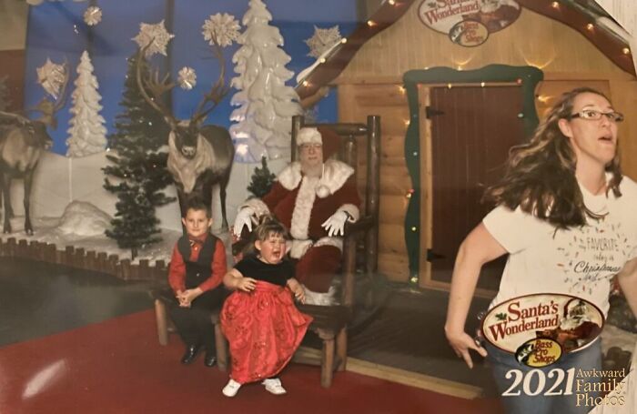 Crying child in red dress sitting next to stoic boy with Santa and reindeer backdrop in awkward family Christmas photo.
