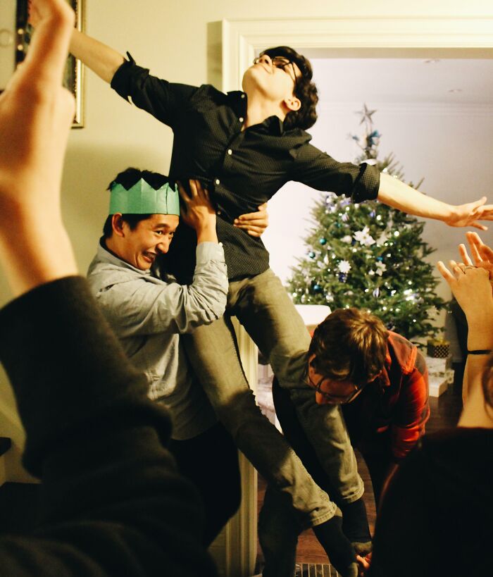 Two men awkwardly lifting a young man in front of a decorated Christmas tree during a funny family Christmas photo.