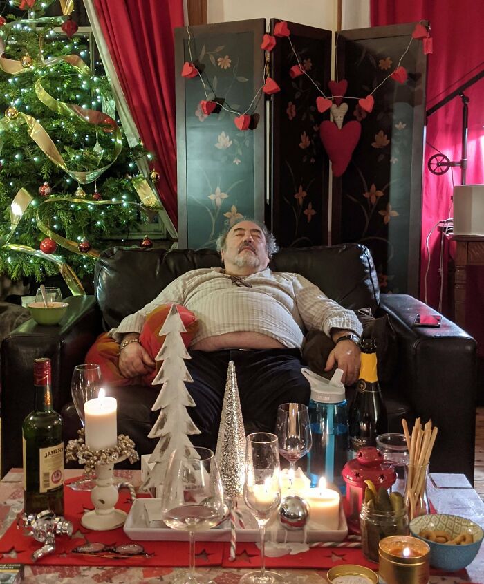 Middle-aged man asleep on couch after family Christmas party with festive decorations and drinks around him, awkward holiday photo.