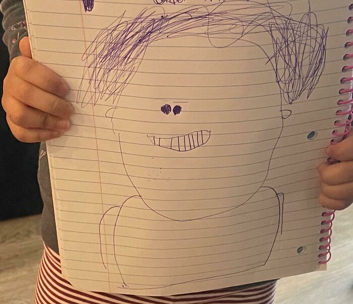 Child holding a funny drawing portrait with no filter, featuring a simple face and scribbled hair on lined notebook paper.