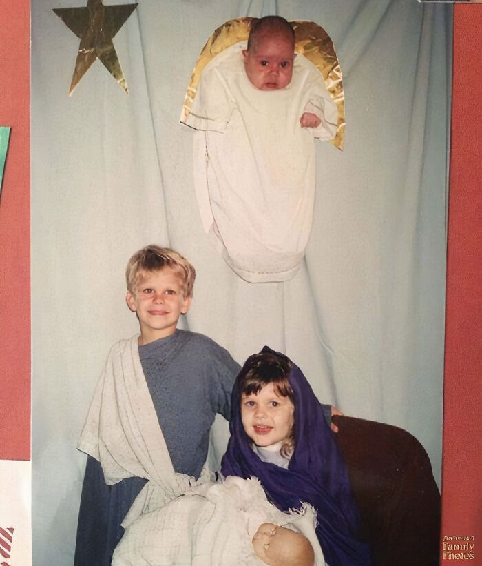 Three children dressed as nativity characters in an awkward family Christmas photo with a baby angel hanging above.