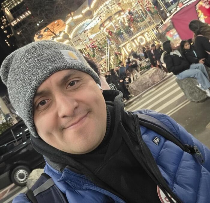 Man wearing a gray beanie and blue jacket smiling at a festive outdoor market with lights and people around. Gifts for the person who has everything.