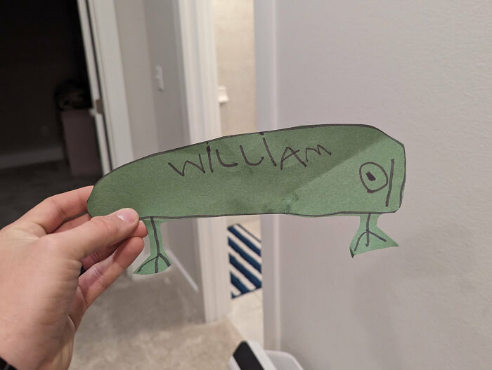 Hand holding a funny kids drawing of a green creature with legs and the name William written on it, a funny drawing portrait.