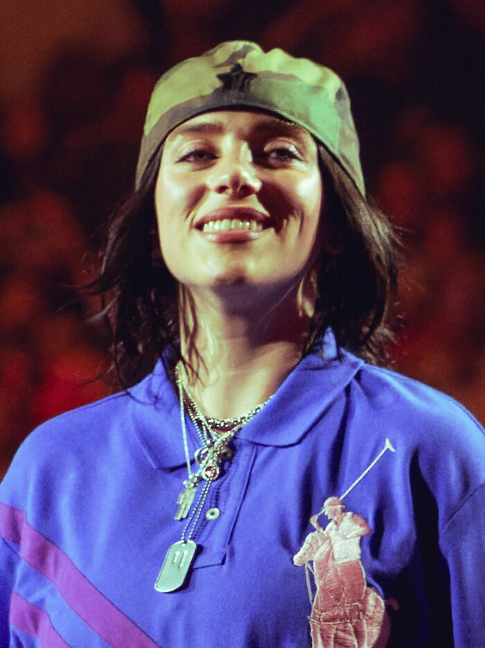 Billie Eilish wearing a green cap and blue shirt with layered necklaces smiling on a dimly lit stage.