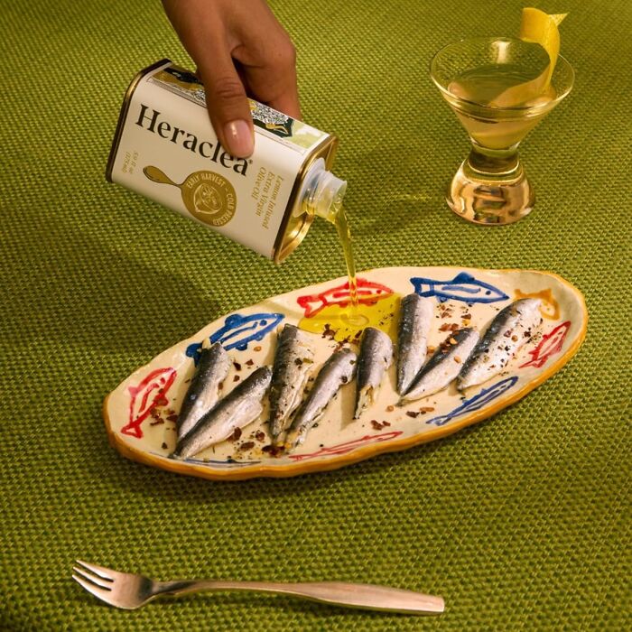 Hand pouring oil over sardines on a colorful fish-themed plate, showcasing unique host gifts for food lovers.