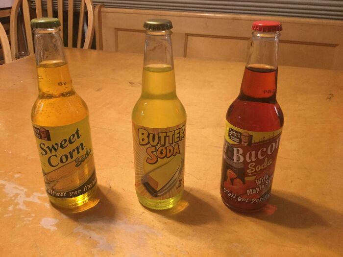 Three bottles of bizarre flavored sodas on a wooden table including sweet corn, butter, and bacon flavors as unusual Christmas gifts.