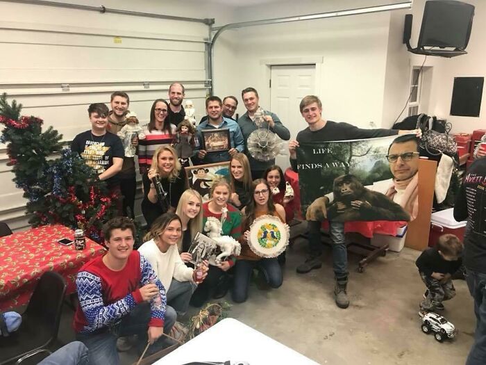 Group of people in a garage holding bizarre Christmas gifts including unusual decorations, photos, and toys.