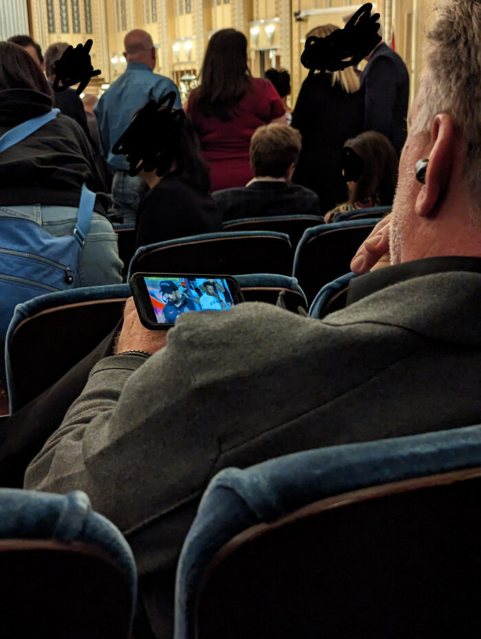 Man in theater watching phone video during ceremony, example of next-level jerks