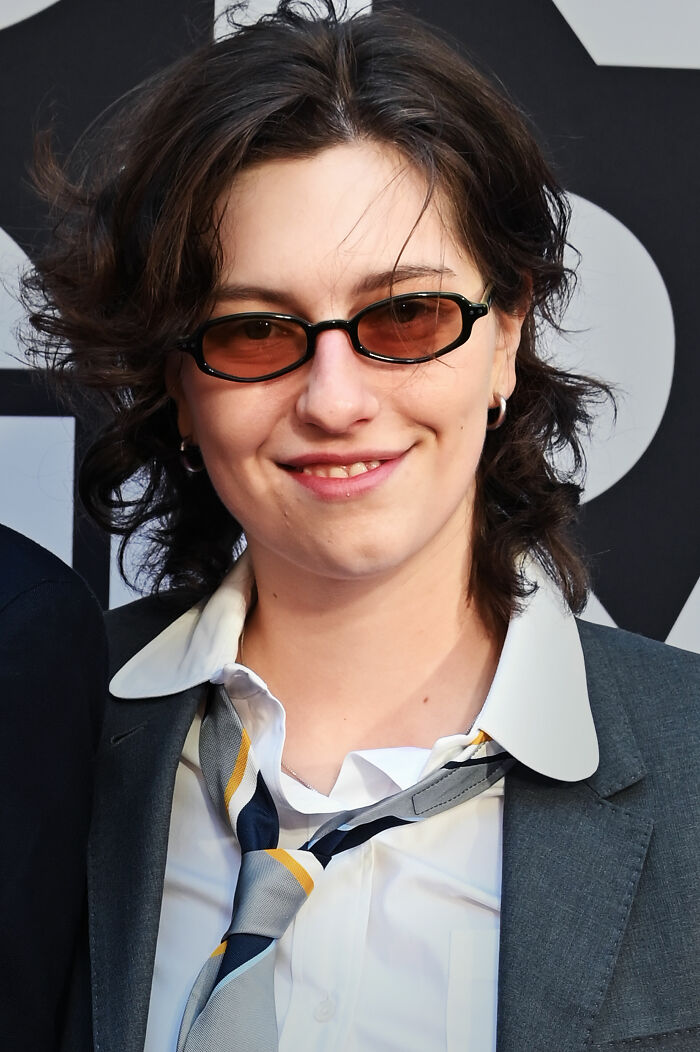 King Princess wearing tinted glasses, a suit jacket, and a loosened tie, smiling at an event.