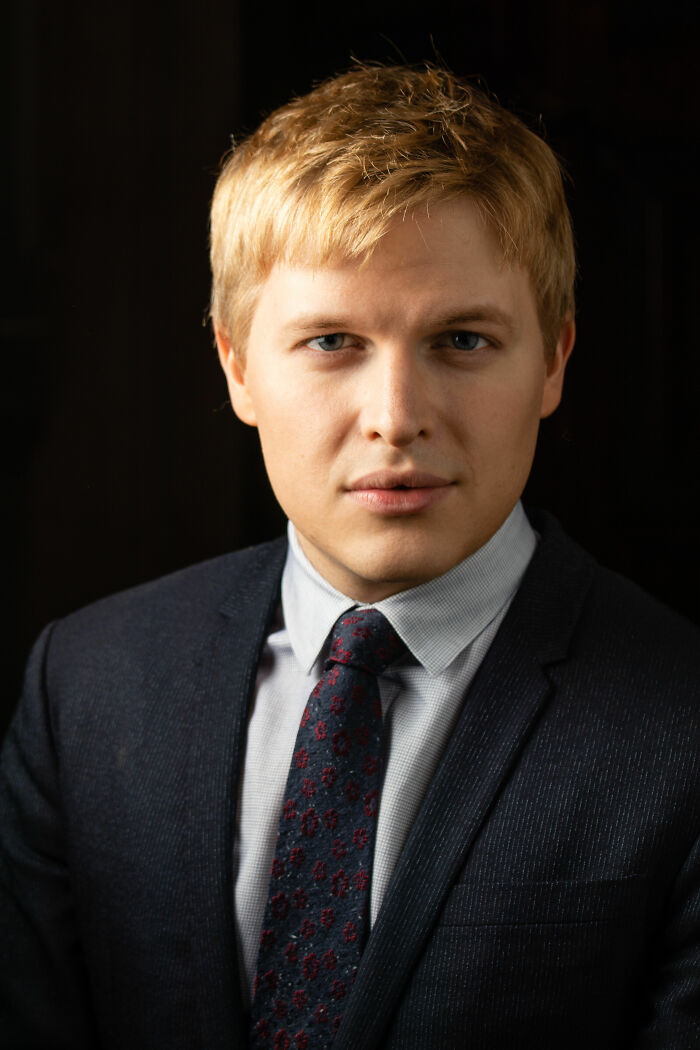 Ronan Farrow wearing a dark suit and patterned tie, posing confidently against a dark background.
