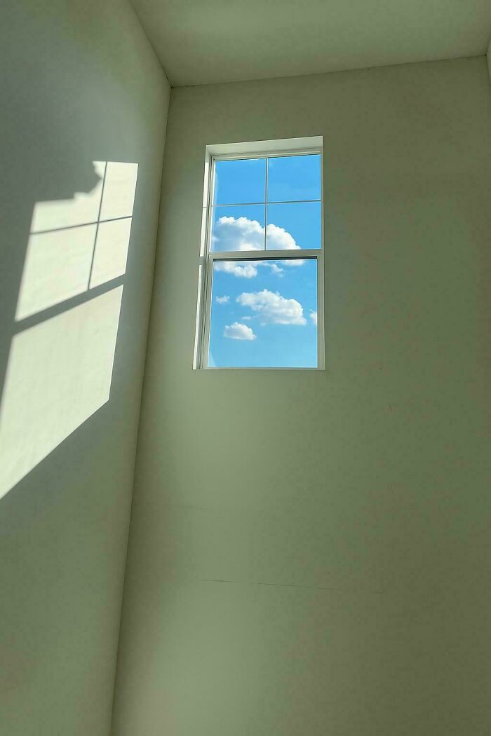 Minimalist liminal space with natural light casting shadows through a high window showing a bright blue sky and clouds.