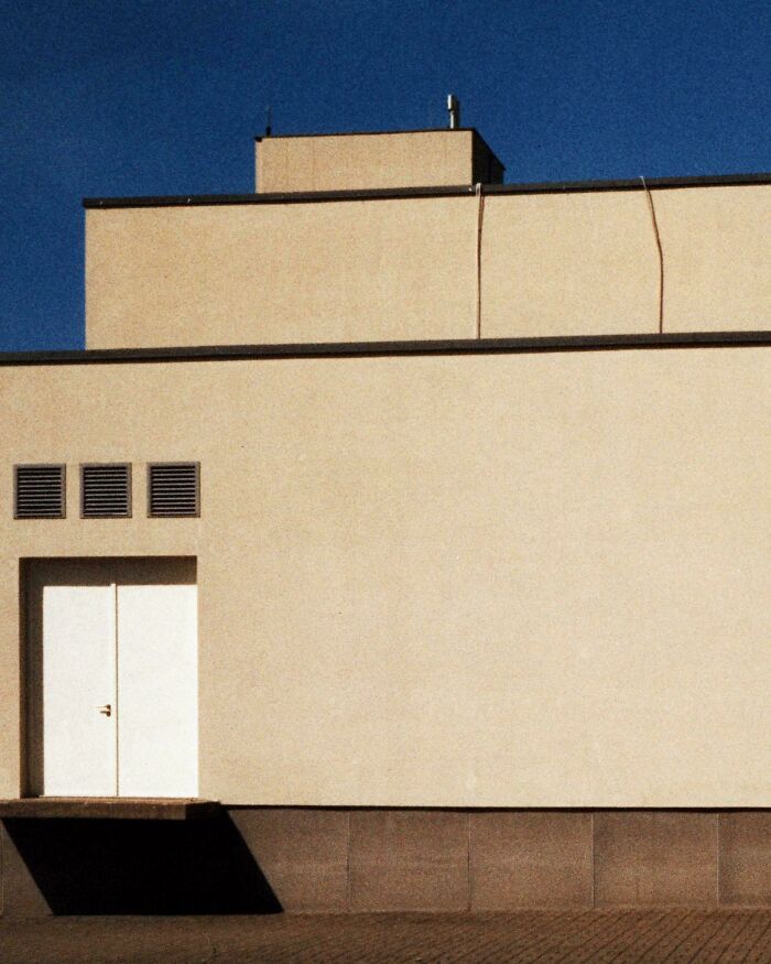 Minimalist beige building exterior with a closed white door under clear blue sky, evoking creepy liminal spaces vibe.