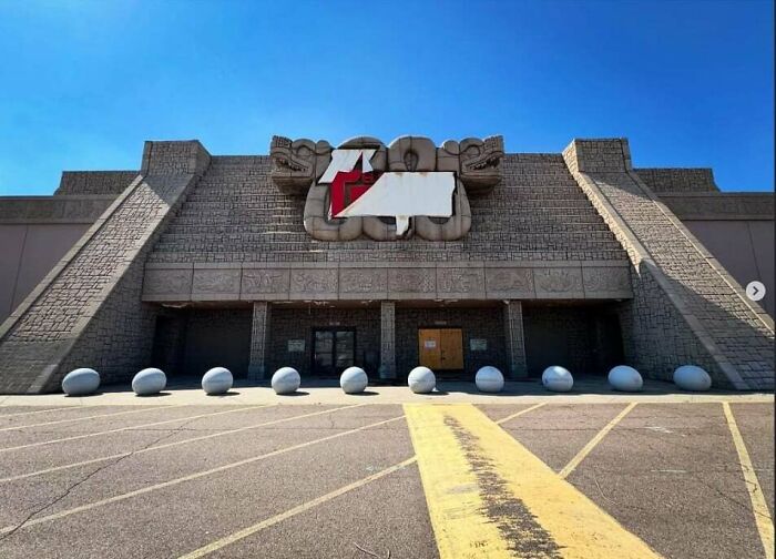 Empty creepy liminal space with stone facade and large blank sign, resembling eerie backrooms atmosphere under clear blue sky.