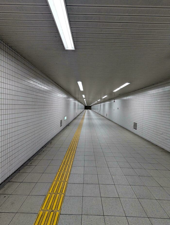 Empty tiled underground passageway with fluorescent lights and a yellow tactile line, evoking creepy liminal spaces.