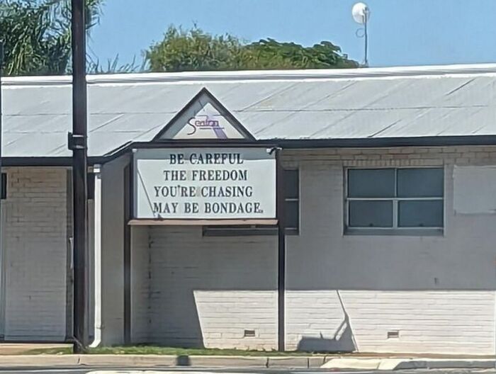 Church sign with a humorous message about freedom and bondage displayed outside a white building under clear skies.