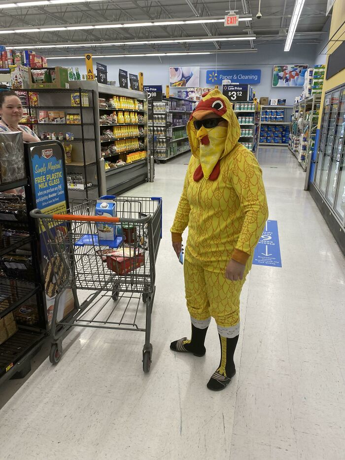 Person dressed in a yellow chicken costume and sunglasses shopping in a Walmart aisle with a cart and groceries.