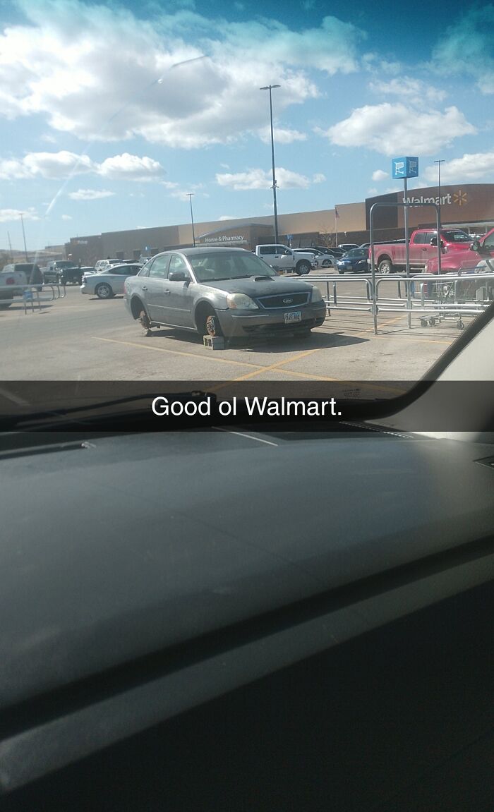Silver car missing wheels parked on blocks in a Walmart parking lot with shopping carts nearby on a sunny day.