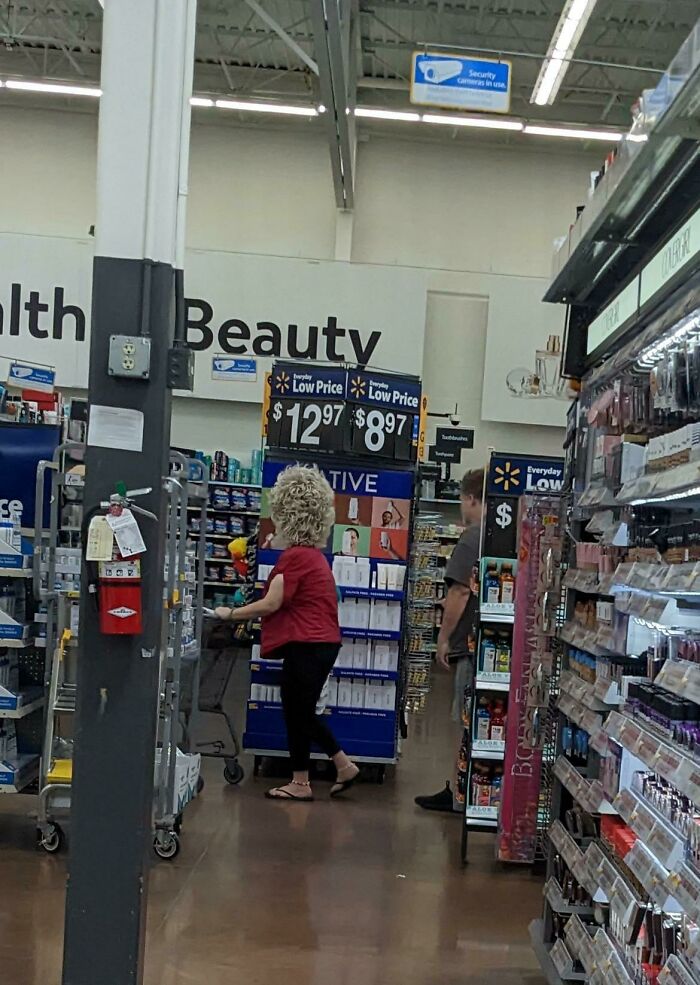 Customer with large curly wig shopping in Walmart beauty aisle near low price signs and shelves of products.
