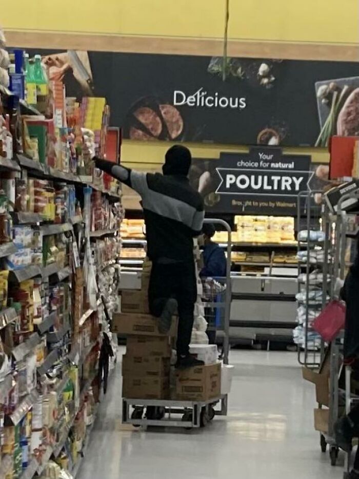 Person standing on boxes stacked on a cart reaching for items on a high shelf inside a Walmart store aisle.