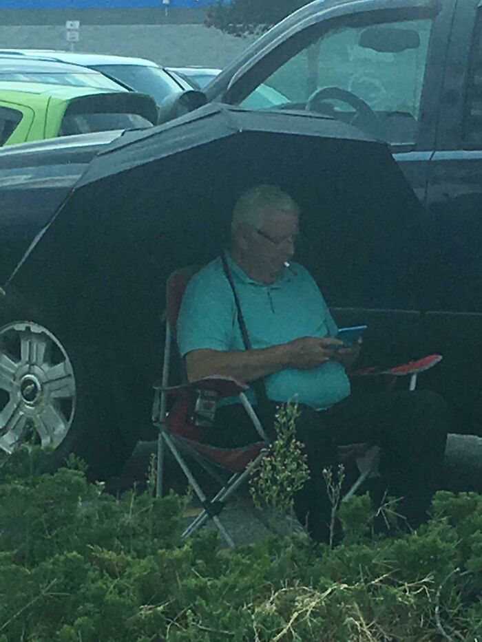 Man seated under a car tire-shaped umbrella in a parking lot, a humorous Walmart people moment captured.