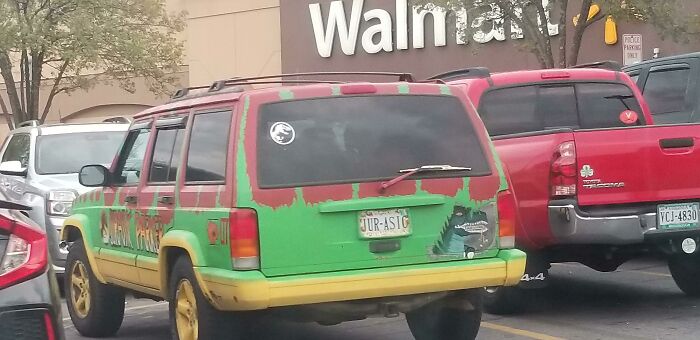 Green and yellow Jeep with a Jurassic Park theme parked in a Walmart parking lot among other vehicles on a cloudy day.