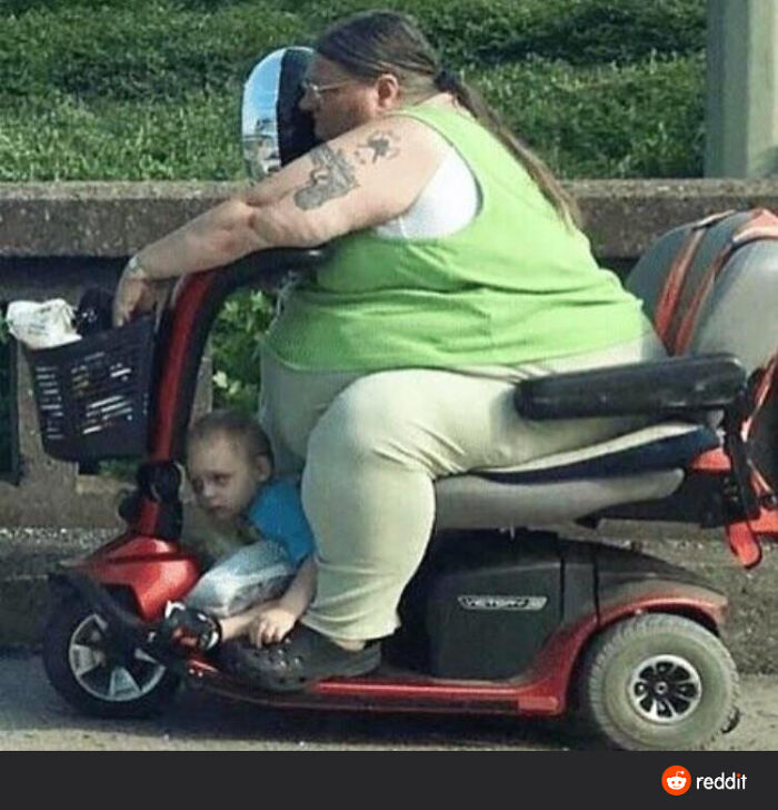 A Walmart person in a green top on a mobility scooter with a child sitting beneath the front footrest.