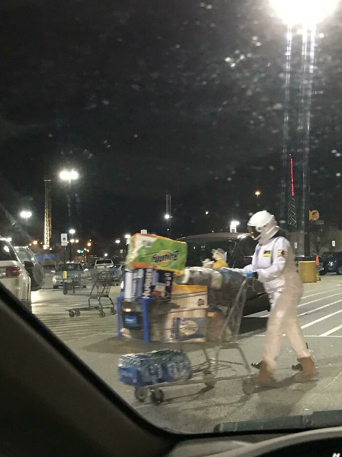 Person dressed as an astronaut pushing a Walmart shopping cart filled with groceries in a parking lot at night.