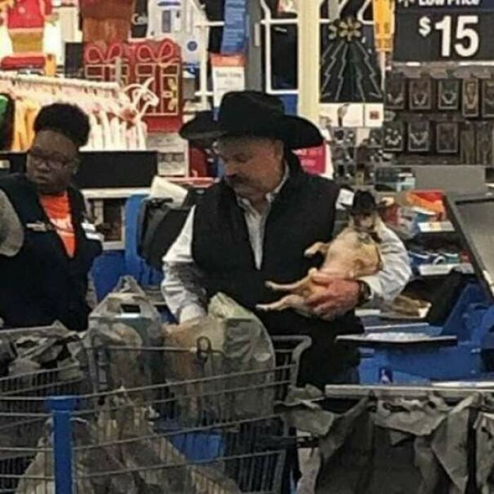 Man wearing cowboy hat holding small dog while checking out at Walmart registers, with Walmart people employee nearby.