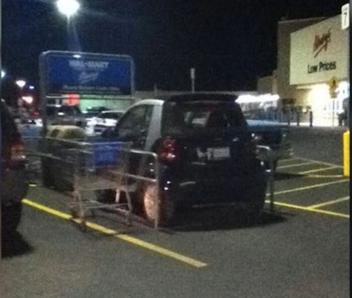 A small black car parked in a Walmart parking lot surrounded by shopping carts in confusing Walmart people scene.