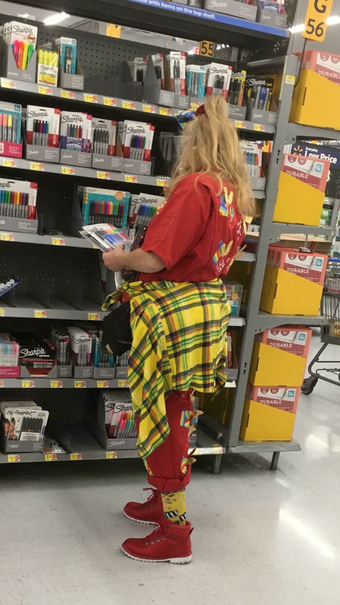 Person in bright red outfit with yellow plaid shirt tied around waist shopping for markers in Walmart aisle.