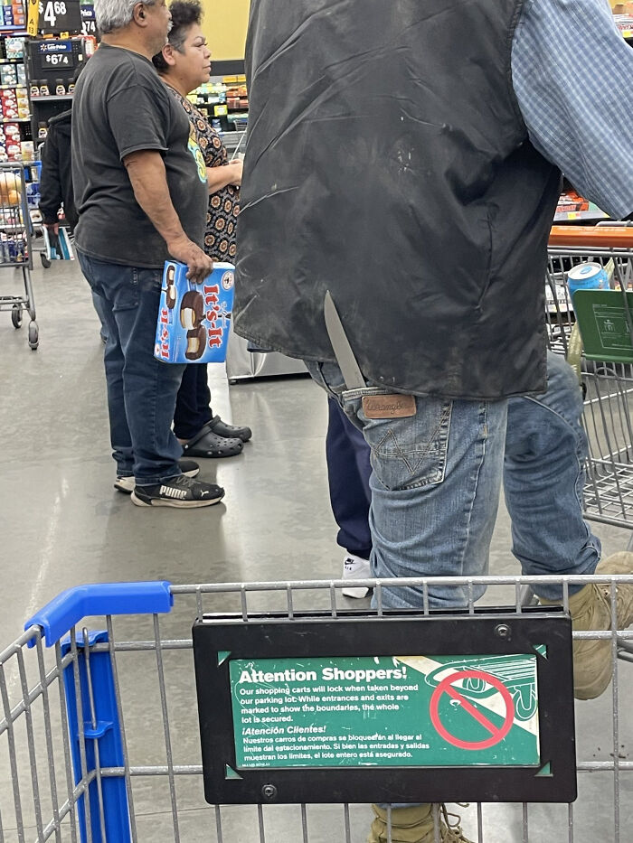 Shoppers in a Walmart aisle with one person wearing a large knife in their back pocket, creating a confusing scene.