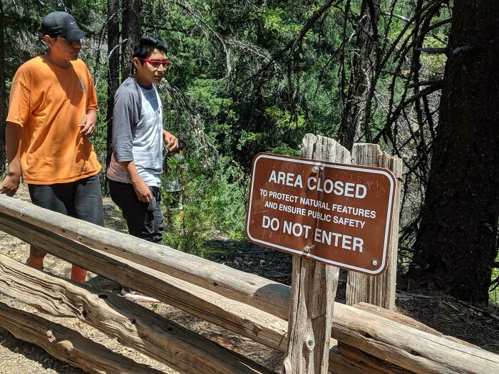 Two hikers ignoring an Area Closed Do Not Enter sign in a forest, example of next-level jerks.