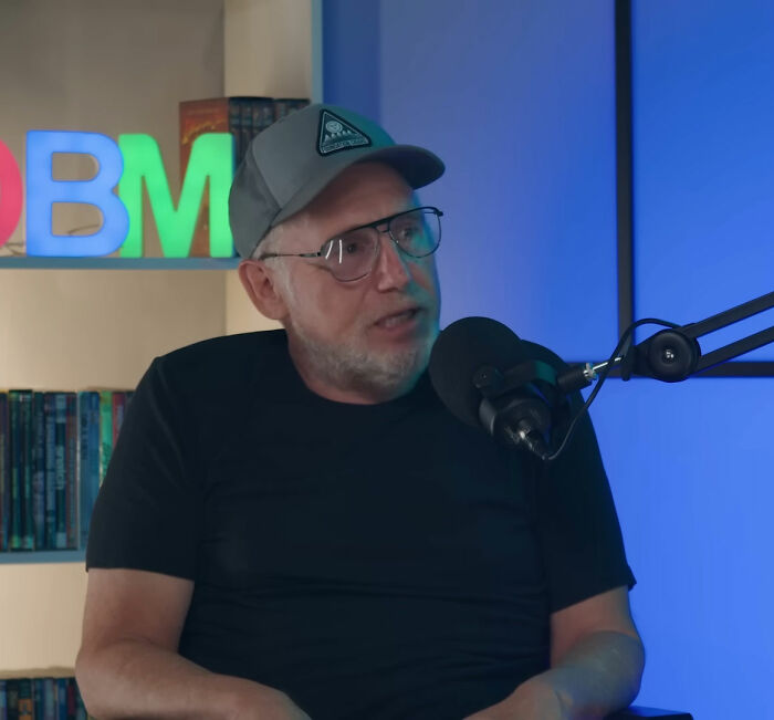 Man wearing glasses and a hat speaking into a microphone during a podcast about Steven Spielberg and Ben Affleck.