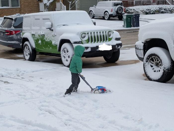 Funny Pictures 2025: child in green hooded jacket pushing a toy snowblower on a snowy driveway with parked cars